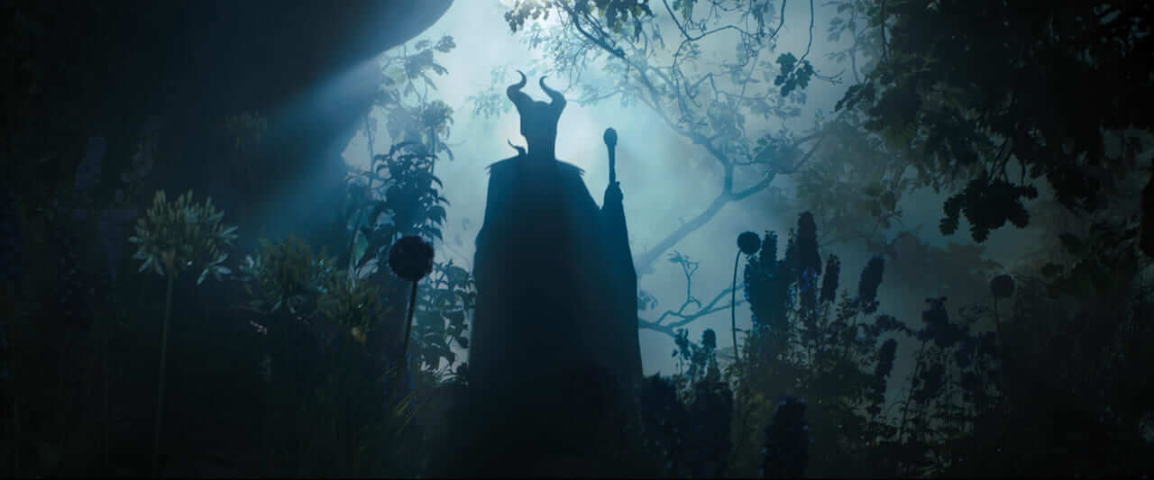 Maleficent in silhouette, horned and wingless, stands with her staff among dark plants in the misty, darkened Moors