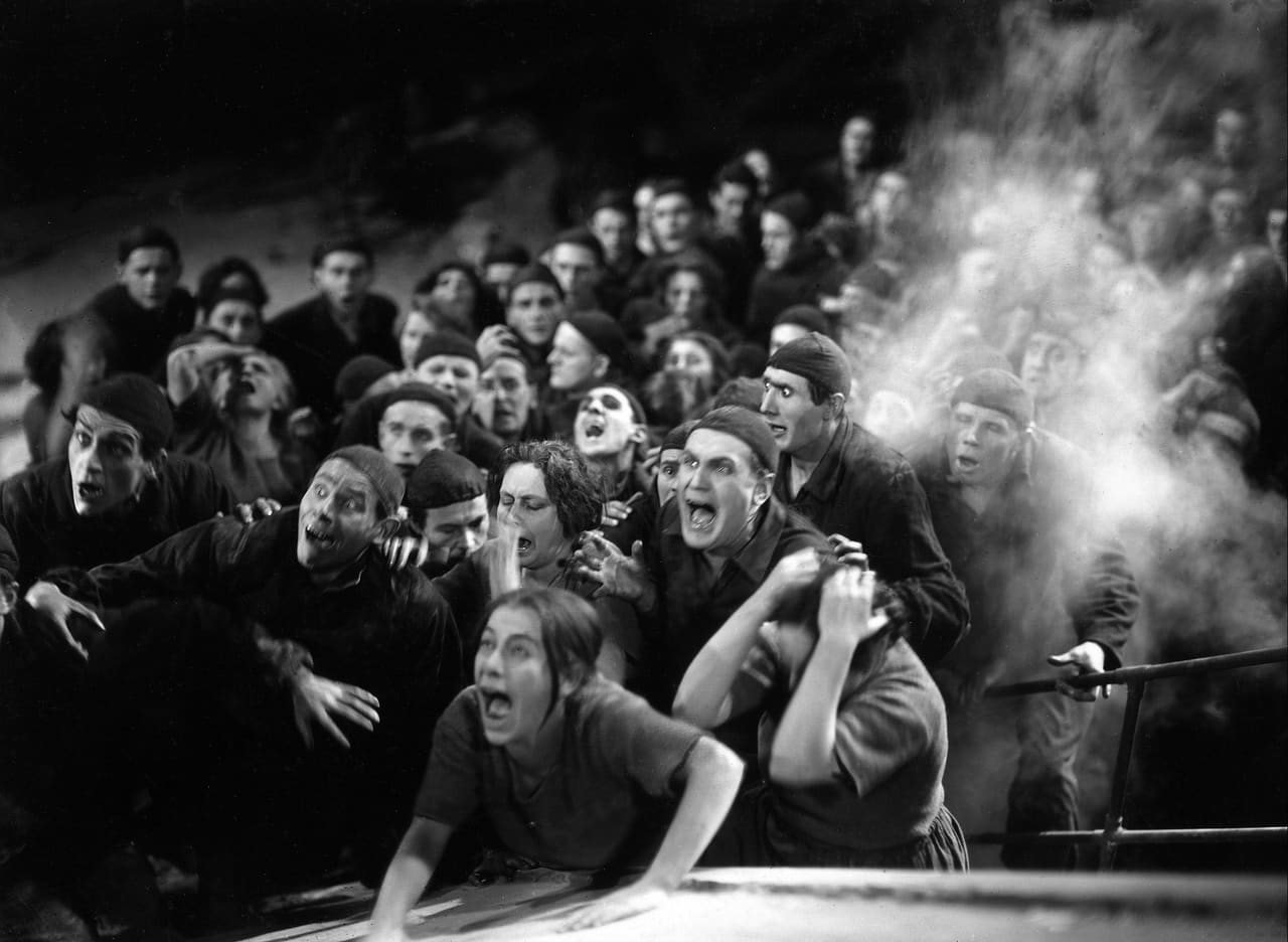 Black-and-white set photograph: a crowd of terrified workers in dark caps and work clothes surges forward, screaming, through clouds of spray