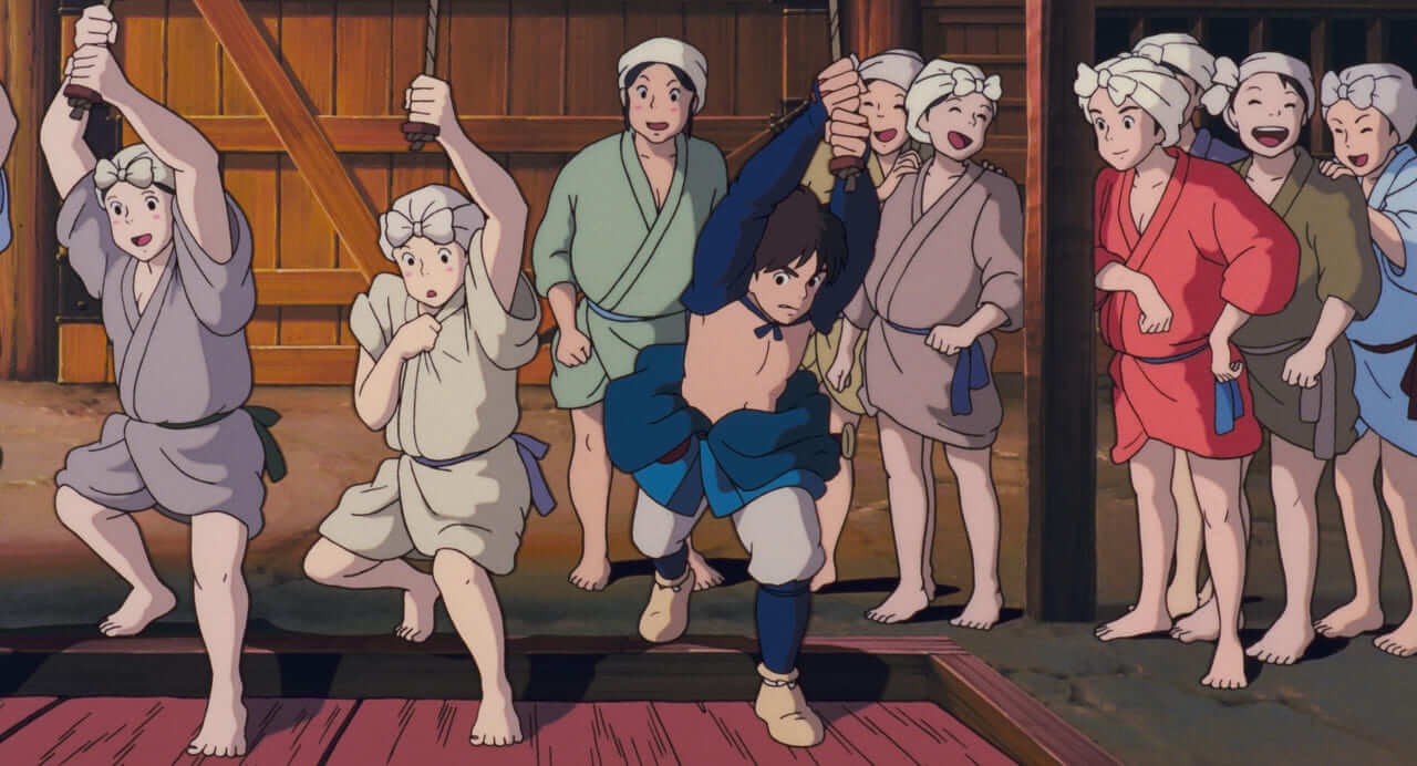 Ashitaka, stripped to the waist, treads Iron Town's great bellows beside two women in work clothes and white headscarves, while more women behind them laugh and cheer him on